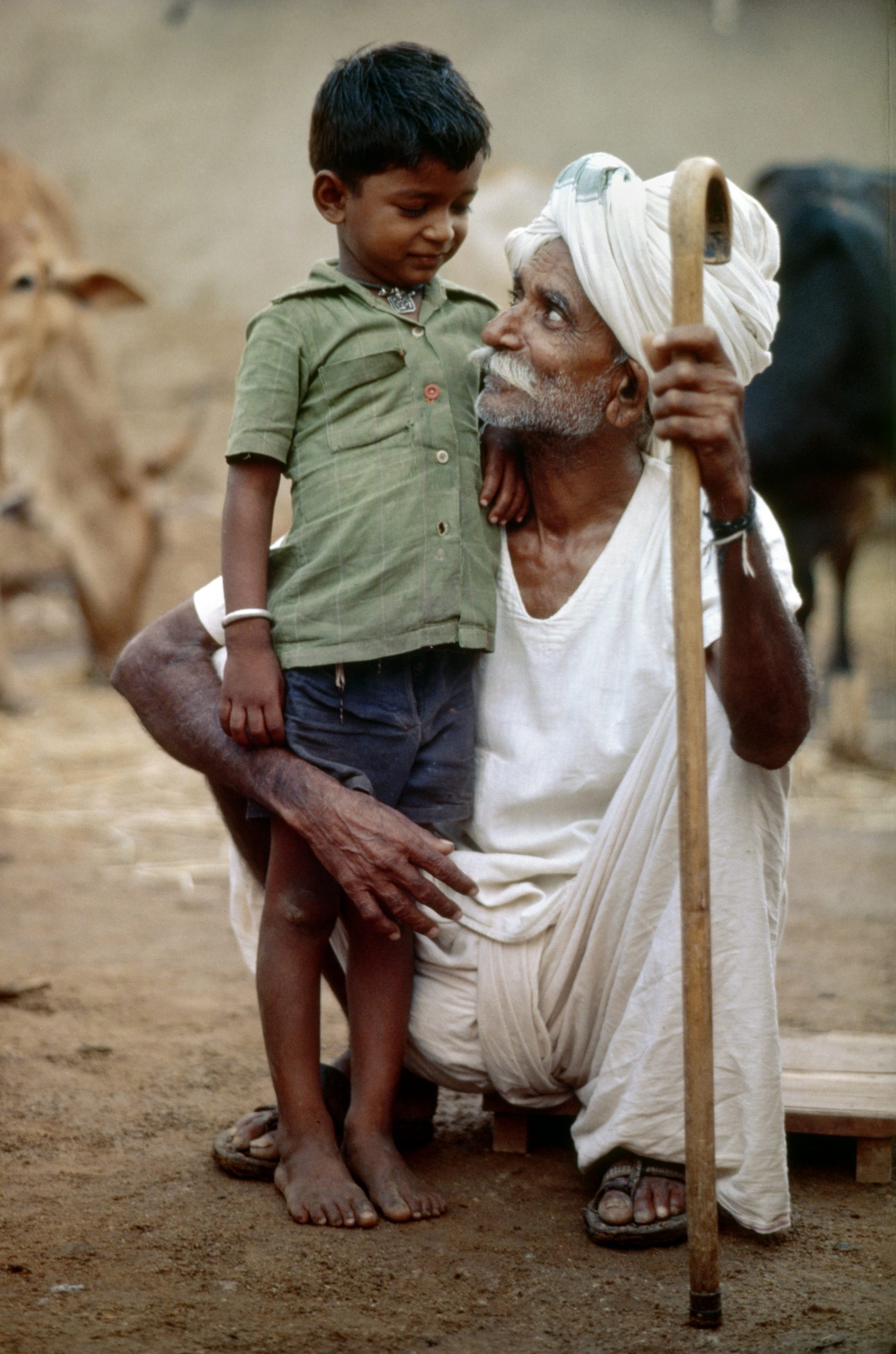A squatting grandfather holding a young boy to his side and making eye contact with him